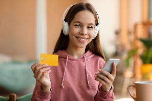 teen girl holding credit card and paying with smartphone, listening music in wireless headphones, sitting at home.