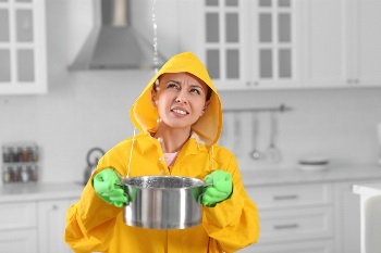 Young woman in raincoat collecting leaking water from ceiling at home.